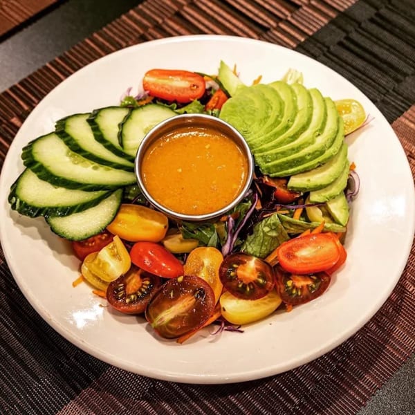 A vibrant hibachi salad with sliced cucumbers, avocado, colorful cherry tomatoes, and a side of dressing.