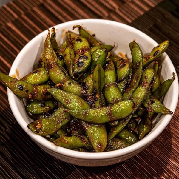 A white bowl filled with grilled edamame, seasoned with spices, on a brown woven placemat.